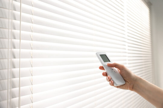 Woman using remote control to adjust window blinds indoors, closeup. Space for text
