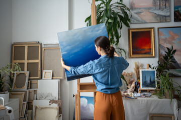 Satisfied female artist holding canvas with artwork of blue sky, putting painting from easel aside,...
