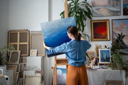 Satisfied female artist holding canvas with artwork of blue sky, putting painting from easel aside, finishing work on picture. Rear view, glad proud woman completing creating masterpiece, piece of art