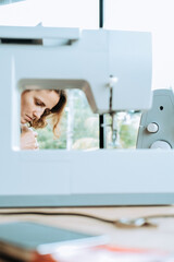 Vertical portrait of a young woman tailor who sits against the background of a sewing machine and sews a dress for a client. Small home sewing business. Concentration on work.