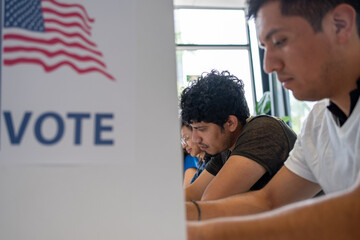 Hispanic young man is seen intently casting his vote in a U.S. election. The voting booth is adorned with an American flag and the word 