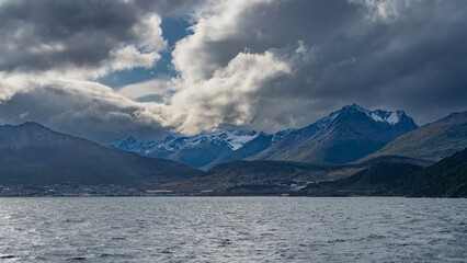 Beautiful snow-capped Andes mountains against the sky and picturesque clouds. The city houses of Ushuaia are visible at the foot. The Beagle Channel is in the foreground. Argentina. Patagonia.