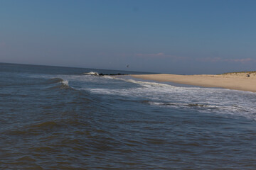 Beautiful image of the Jersey shore. The white froth of the waves crashing in to the beautiful brown sand of the beach. A small jetty with black rocks can be seen. The blue sky has some white clouds.