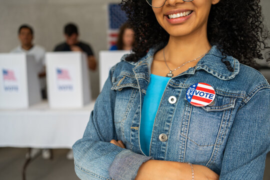 close up on a 2024 vote pin button of a young African American woman smiling at a US election polling station. In the background a diverse group of people votes in voting booths, democracy in America