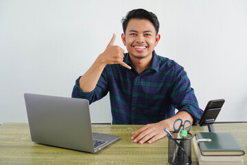 young asian man sit work at wooden desk with pc laptop. Achievement business career lifestyle concept. doing phone gesture like says call me back