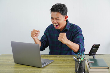 Happy young asian man sit work at wooden desk with pc laptop. Achievement business career lifestyle concept. doing winner gesture while looking at the laptop screen