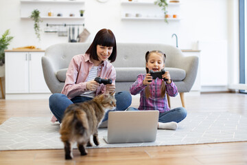 Mother and daughter enjoying leisure time playing video games in cozy living room. Both holding controllers, smiling, and sitting on floor with laptop. Pet cat walking around adding warmth to scene.