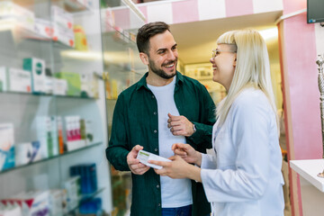 Cheerful pharmacist chemist woman giving vitamins, antibiotics to young man in modern pharmacy.
