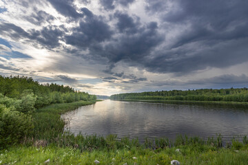 A peaceful river with green trees along the banks, reflecting gray and white clouds. The setting suns golden light casts a warm glow over the scene.