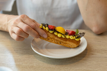 A hand reaches for a delicious eclair topped with fresh fruit, chocolate sprinkles, and a swirl of...