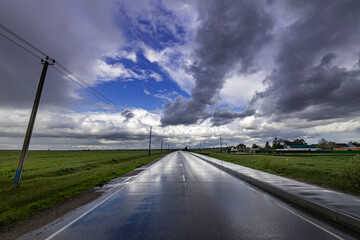 A wet asphalt road cuts through a green field, disappearing into the distance under a sky filled with dramatic, grey clouds that threaten more rain..