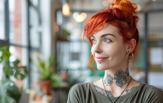 A smiling woman with red hair and tattoos looks out a window while sitting in an office