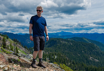 Adventurous athletic male hiker standing on a mountain top on a sunny day with rugged mountains in the background in the Pacific Northwest.