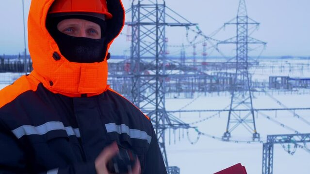 Engineer in winter gear is at electricity substation, equipped with walkie talkie for communication. Setting is cold with snow, and worker is clad in protective gear. Work in industry, at power plant