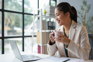 Obraz premium Attractive businesswoman holding cup of hot drink and looking away with smile while sitting in office.