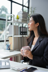 Fototapeta premium Asian businesswoman sitting, relaxing, holding a hot coffee mug, working with laptop on table, online finance, documents, taxes, reports, marketing, statistics, business analysis research concept.