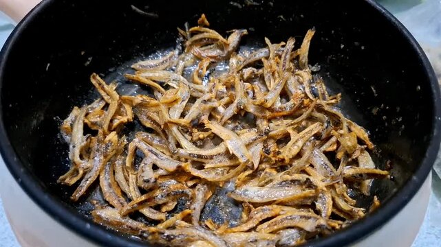 footage of frying salted fish or anchovies using an electric pot and frying with a tablespoon. Cooking quickly so as not to burn and smell of smoke.