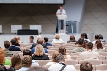 Group of business professionals sitting in an auditorium, attending a presentation during a tech conference. The speaker is standing at a podium.