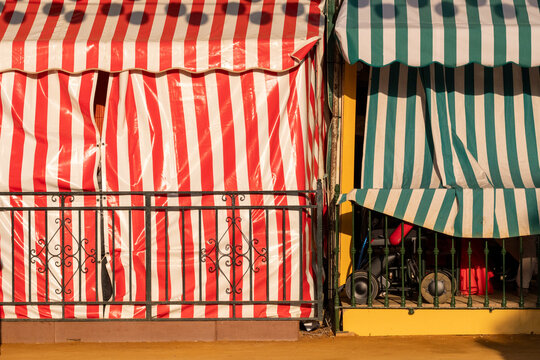 Vivid red and green striped awnings providing shade over a patio with iron railings in the foreground, evoking a sense of vibrant European street market ambiance in Seville