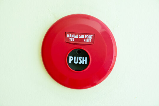 The image shows a close-up of a circular, bright red manual fire alarm call point mounted on a light-colored wall.