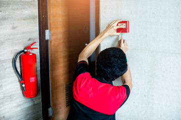 Technician testing red fire alarm system call point in office building corridor next to fire extinguisher. © Koonsiri