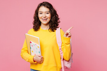 Young smiling happy teen girl student she wear yellow casual clothes backpack bag hold books point finger aside isolated on plain pastel light pink background. High school university college concept.