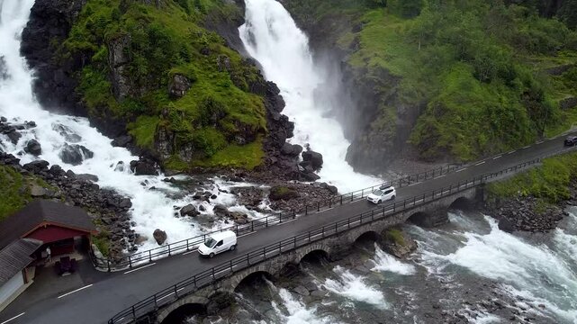 Drone footage of L&aring;tefossen Waterfall cascading beside a stone bridge in Norway. Cars pass over the bridge, enhancing the scenic and powerful natural beauty.