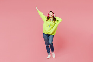Full body young smiling happy cheerful ginger woman she wear green hoody casual clothes listen to music in headphones isolated on plain pastel light pink background studio portrait. Lifestyle concept.