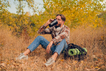 Bearded adult man sitting at a rest stop during a hike, drinking hot coffee from a thermos, enjoying the tranquility of the forest.