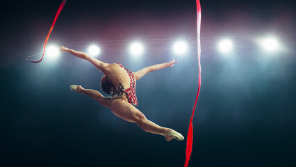Female Gymnast in Mid-air, Executing Flawless Split Leap With Ribbon Under Bright Stadium Lights....