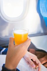 View of window seat on board of airplane with a cup of orange juice in woman’s hand.
