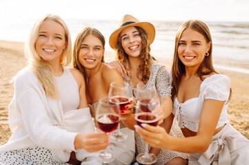 The company of gorgeous female friends having fun, cheers and drink wine, and enjoy picnic on the beach. Hen party.