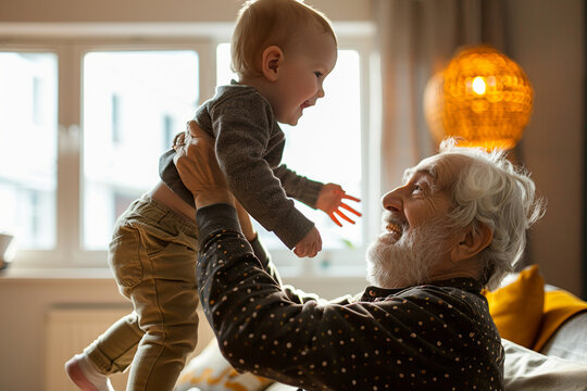 An elderly man lifting his joyful grandchild in the air inside a cozy, warmly lit living room.