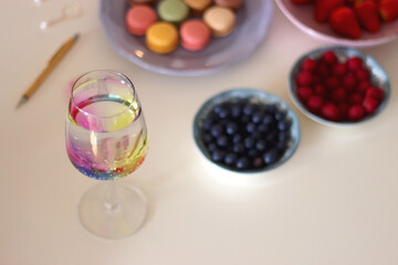 Plate of pastel macarons, cookies and chocolate, cup of tea of coffee, glass of bubble water, various berries, books and accessories on the table. Selective focus, pastel colors.