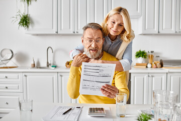 A man tenderly holds a woman above a table in a cozy kitchen, a scene of affection and closeness between a mature couple.