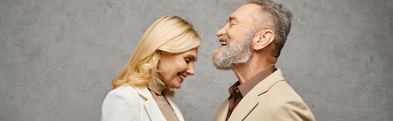 An elegant, mature loving couple in debonair attires pose together on a gray backdrop.