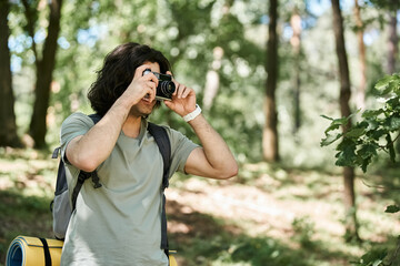A young man hikes through the woods on a sunny summer day, capturing the beauty of nature with his camera.