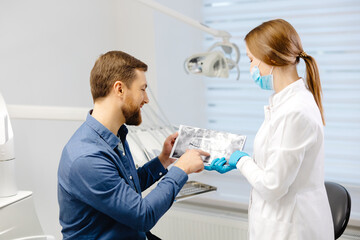 Young attractive man visiting dentist, sitting in dental chair at modern light clinic. Young woman dentist holding x ray image.