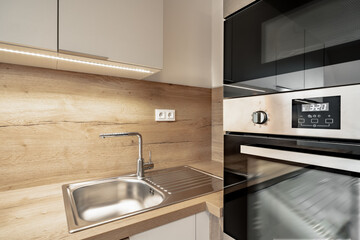 
A modern kitchen detail with a stainless steel sink, wooden backsplash, white cabinets, under-cabinet lighting, and built-in black oven.