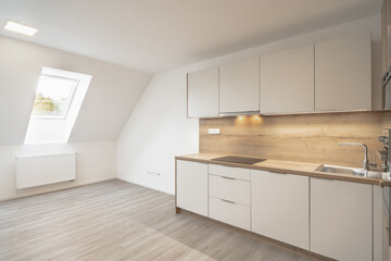 
A modern, bright kitchen with a slanted ceiling, skylight window, white cabinets, wooden countertops, light wooden flooring, and a radiator.