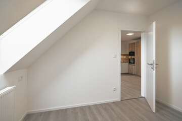 A bright, minimalist room with a slanted ceiling, white walls, a radiator, and an open door leading to a modern kitchen with wooden cabinets and appliances.