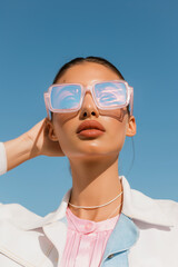 Fototapeta premium Woman wearing holographic sunglasses and pink shirt, posing against blue pastel background.