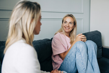 Two happy smiling women friends talking while sitting on the sofa in a cozy living room.