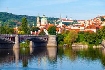 A stone bridge spans the Vltava River in Prague, Czechia. Red-roofed buildings line the hills in the background, with a church spire visible.