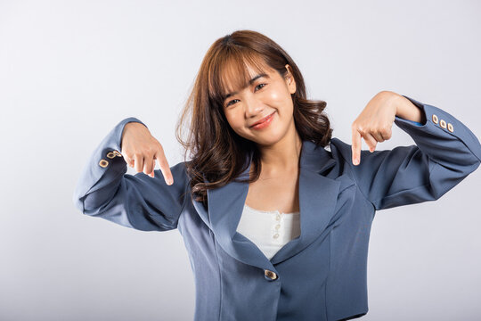 An Asian business woman stands in a studio portrait, smiling and making a two-finger pointing gesture downwards. Isolated on white background, ideal for advertisements and promotions