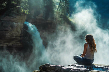 Obraz premium A woman sitting on a rock by a waterfall, with mist rising around her.