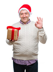 Handsome senior man wearing christmas hat and holding gift over isolated background doing ok sign with fingers, excellent symbol