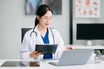 Confident young Asian female doctor in white medical uniform sit at desk working on computer. Smiling use laptop write in medical journal