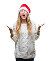 Young beautiful blonde woman wearing christmas hat over isolated background crazy and mad shouting and yelling with aggressive expression and arms raised. Frustration concept.