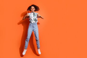 Full body photo of american woman in jeans and summer stylish shirt jumping trampoline thumbs up recommendation isolated on orange color background